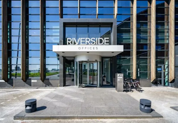 Ingang van het Riverside Offices-gebouw aan de Rivium Promenade met een glazen gevel en draaideur.